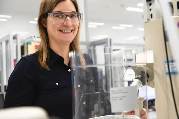 Femme souriante portant des lunettes de sécurité dans un laboratoire. Elle est devant un équipement scientifique, avec un environnement de travail moderne et organisé en arrière-plan.