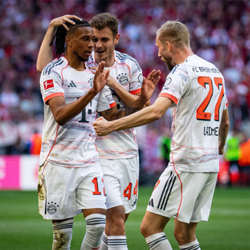 Trois joueurs de football de l'équipe du Bayern Munich se réjouissent après un but. L'un des joueurs reçoit des félicitations de ses coéquipiers, montrant un moment de camaraderie et d'excitation. Les maillots sont blancs avec des touches de couleur.