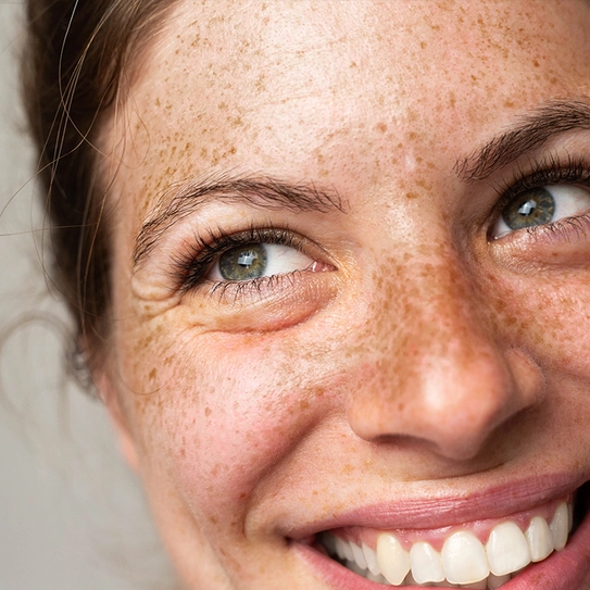 A close-up image of a smiling woman with brown hair, freckled skin, and bright green eyes. Her expression conveys happiness and warmth, highlighting her cheerful demeanor.