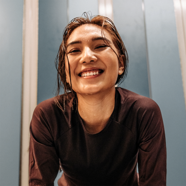 Une jeune femme souriante avec les cheveux mouillés, portant un haut à manches longues sombre. Elle est penchée en avant, avec un fond bleu. Son expression dégage de la joie et de la satisfaction.
