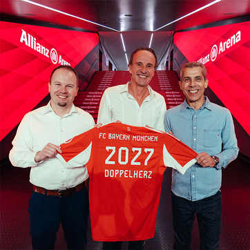 Trois hommes se tiennent dans le couloir de l'Allianz Arena, tenant un maillot de football rouge du FC Bayern Munich. Le maillot affiche le numéro 2027 et le mot "DOPPELHERZ". L'ambiance est chaleureuse et festive, reflétant une annonce importante pour le club.