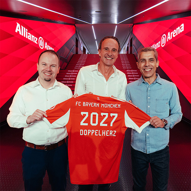 Trois hommes se tiennent dans le couloir de l'Allianz Arena, tenant un maillot de football rouge du FC Bayern Munich. Le maillot affiche le numéro 2027 et le mot "DOPPELHERZ". L'ambiance est chaleureuse et festive, reflétant une annonce importante pour le club.
