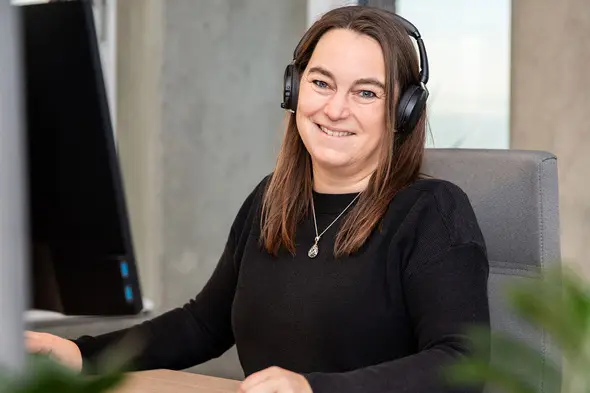 Femme souriante portant des écouteurs, assise devant un bureau avec un ordinateur. Elle a les cheveux bruns et longs, et porte un pull noir. En arrière-plan, on aperçoit une fenêtre et une plante.