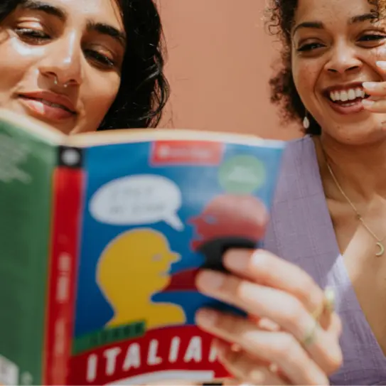 Two women are engaged in conversation while looking at a book titled "Learn Italian." One woman has dark hair and a septum ring, while the other has curly hair and appears to be smiling. The background is a soft, warm color.