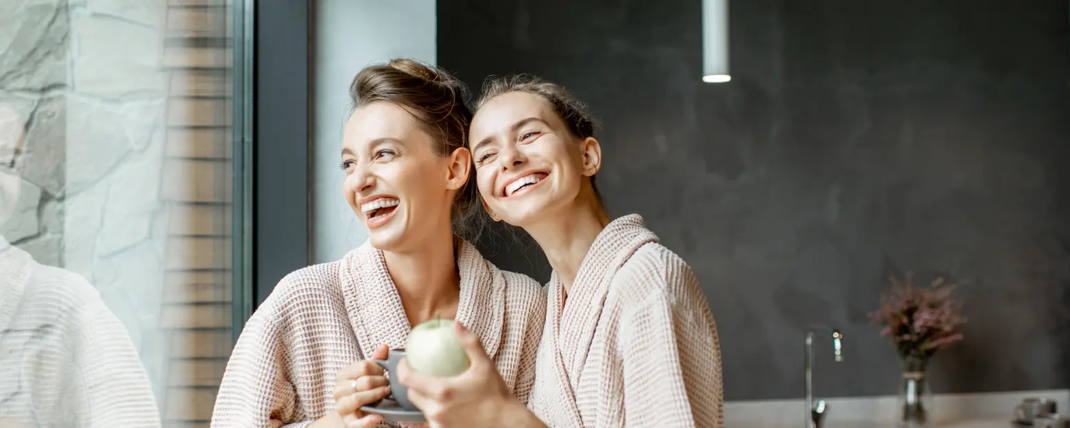 Two women in cozy, matching bathrobes stand together by a large window, smiling and laughing. One holds a cup, while the other cradles a green apple. They appear relaxed and joyful, enjoying each other's company in a modern kitchen setting.
