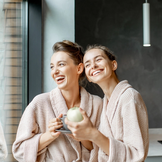 Two women in cozy, matching bathrobes stand together by a large window, smiling and laughing. One holds a cup, while the other cradles a green apple. They appear relaxed and joyful, enjoying each other's company in a modern kitchen setting.