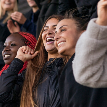Trois femmes souriantes sont ensemble dans une ambiance festive, levant les bras et affichant une grande joie. Elles portent des vêtements d'extérieur sombres. En arrière-plan, d'autres personnes semblent également apprécier le moment. L'image transmet un sentiment de camaraderie et de célébration.